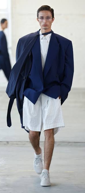 Man walking down a runway with a navy blue top, and while under shirt and jeans and glasses.
