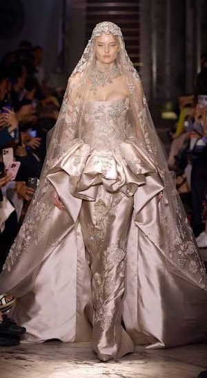 lady waliing down a runway at a fashion show while wearing a while dress with a block bottom.